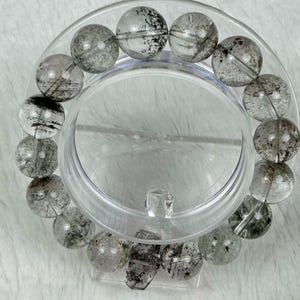May include: A clear quartz bracelet with black inclusions, displayed on a clear plastic stand. The bracelet features large, round beads with visible internal patterns. The beads are arranged in a circular shape, showcasing the natural variations within the quartz.