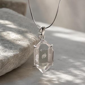Puede incluir: Un colgante de cristal de cuarzo transparente, hexagonal y de doble terminación, suspendido de una fina cadena de serpiente plateada. El cristal está sujeto por un engaste metálico de cuatro puntas, mostrando su estructura natural y sus inclusiones.