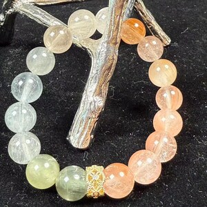 Puede incluir: Una pulsera de cuentas con un degradado de piedras esféricas translúcidas que pasan de tonos verde pálido y transparentes a tonos melocotón y naranja cálidos. La pulsera incluye una cuenta espaciadora de metal dorado con filigrana.
