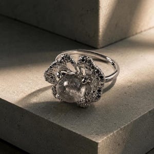 May include: A silver-toned ring featuring a central spherical clear gemstone with visible internal inclusions. The setting is shaped like an open flower with textured petals accented by small, dark, round stones embedded along the edges.