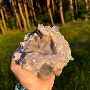 May include: A large, irregularly shaped, gray rock with a rough, textured surface. The rock is held in a hand, with a green and white braided bracelet visible on the wrist. The background features a blurred view of trees and grass.
