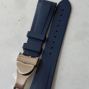 Puede incluir: Una correa de reloj azul marino con un cierre de acero inoxidable. El cierre está grabado con la palabra "TUDOR". La correa está hecha de un material flexible y tiene varios agujeros para ajustar.