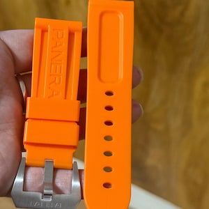 Könnte beinhalten: Hellorangefarbenes Uhrenarmband mit dem eingeprägten Markennamen "PANERAI". Das Armband hat eine silberfarbene Schnalle und besteht aus flexiblem Material. Das Armband ist für eine Uhr konzipiert.