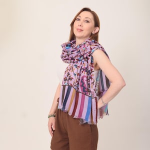 May include: A woman wearing a colorful scarf with a floral pattern and striped border. The scarf is draped over a white top and paired with brown shorts. The scarf's colors include purple, blue, and pink.