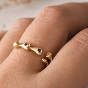 May include: A gold open ring featuring an organic, fluid shape with a small embedded red gemstone and a small embedded dark blue gemstone. The ring has a polished finish and a gap between the two ends, which are rounded and smooth in texture.