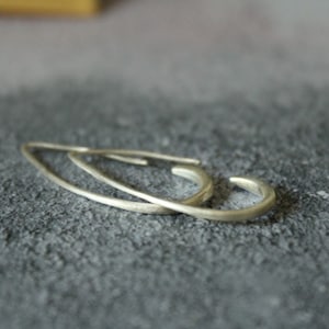 May include: A pair of silver-colored, minimalist hoop earrings. The earrings have a simple, curved design with a smooth, matte finish. The earrings are displayed on a textured, gray surface.