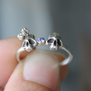 May include: A silver ring with two skull-shaped charms and a small blue gemstone in the center.