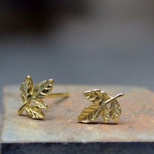 Puede incluir: Un par de pendientes de botón de hoja de oro. Los pendientes son pequeños y delicados, con dos hojas en cada pendiente.