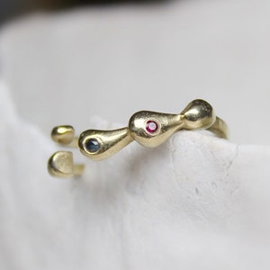 Może przedstawiać: Złoty pierścionek z trzema kamieniami w kształcie łzy. Kamienie są osadzone w pierścionku i są ciemnoniebieskie, czerwone i ciemnoniebieskie.