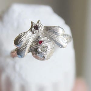 May include: A silver-colored bee ring with a detailed design. The ring features a bee with wings and a body adorned with two small red gemstones. The ring is set against a white background.