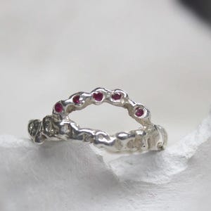 Op de afbeelding: Een zilveren ring met een textuurontwerp, bezet met kleine rode edelstenen. De ring wordt gepresenteerd op een wit, getextureerd oppervlak, dat de ingewikkelde details en het vakmanschap laat zien. De edelstenen zijn gelijkmatig verdeeld.