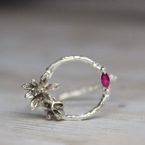 Può includere: Un anello d'argento con un design floreale e una piccola gemma rossa di forma ovale. L'anello ha una fascia strutturata e un'estetica delicata ispirata alla natura. L'anello è posto su uno sfondo sfocato.