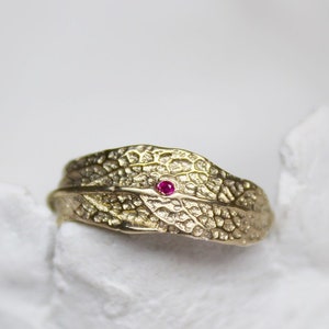 May include: A gold ring with a textured leaf design and a small red gemstone in the center.