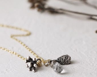 May include: A delicate gold chain necklace featuring two distinct charms: a small, dark, textured, multi-pronged metal piece and a second charm consisting of a clear, faceted, teardrop-shaped stone paired with a small, dark, textured metal element.