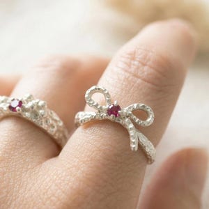 May include: Two silver rings with red gemstones. One ring has a textured band with a cluster of small details and a gemstone. The other ring is shaped like a bow with a gemstone in the center. The rings are on a finger.