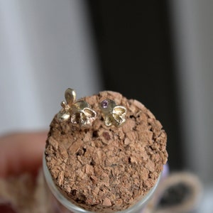 Peut inclure: Deux boucles d'oreilles en forme de cr&acirc;ne en or avec une petite pierre pr&eacute;cieuse violette dans chaque &oelig;il. Les boucles d'oreilles sont pr&eacute;sent&eacute;es sur une surface en li&egrave;ge.