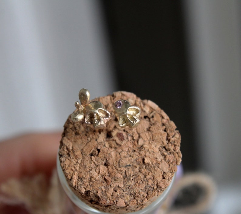 Peut inclure: Deux boucles d'oreilles en forme de cr&acirc;ne en or avec une petite pierre pr&eacute;cieuse violette dans chaque &oelig;il. Les boucles d'oreilles sont pr&eacute;sent&eacute;es sur une surface en li&egrave;ge.