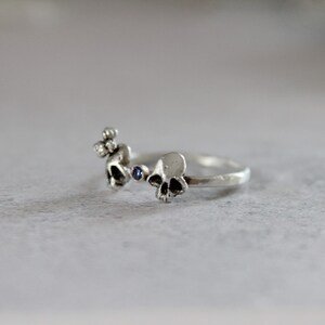 May include: A silver ring with three skull designs and a blue gemstone. The ring is made of a textured metal and has a simple, minimalist design.