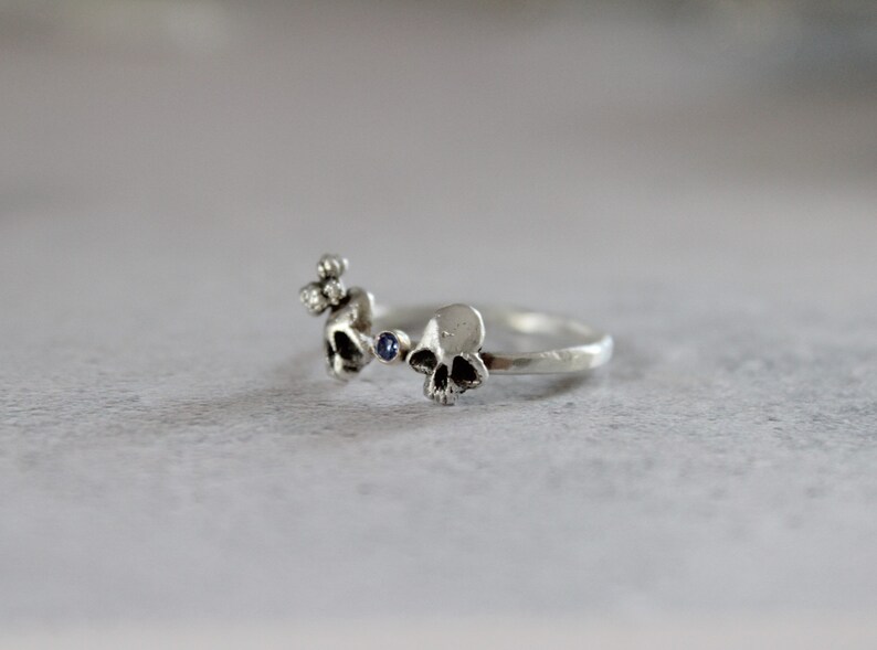 May include: A silver ring with three skull designs and a blue gemstone. The ring is made of a textured metal and has a simple, minimalist design.