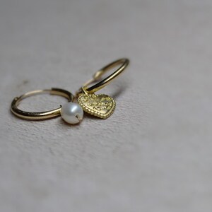 May include: Gold hoop earrings with a small pearl charm and a heart charm. The heart charm has a textured surface.