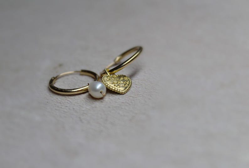 May include: Gold hoop earrings with a small pearl charm and a heart charm. The heart charm has a textured surface.
