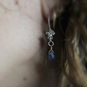 May include: A silver skull charm earring with a blue gemstone dangle. The skull is adorned with small flower details.
