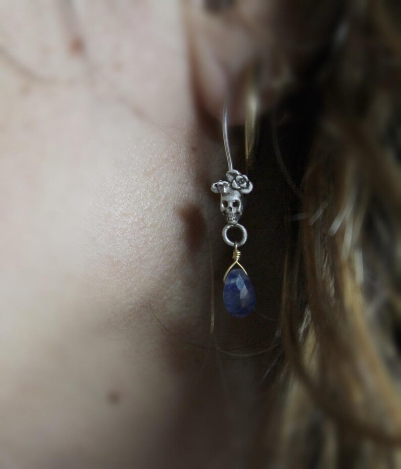 May include: A silver skull charm earring with a blue gemstone dangle. The skull is adorned with small flower details.