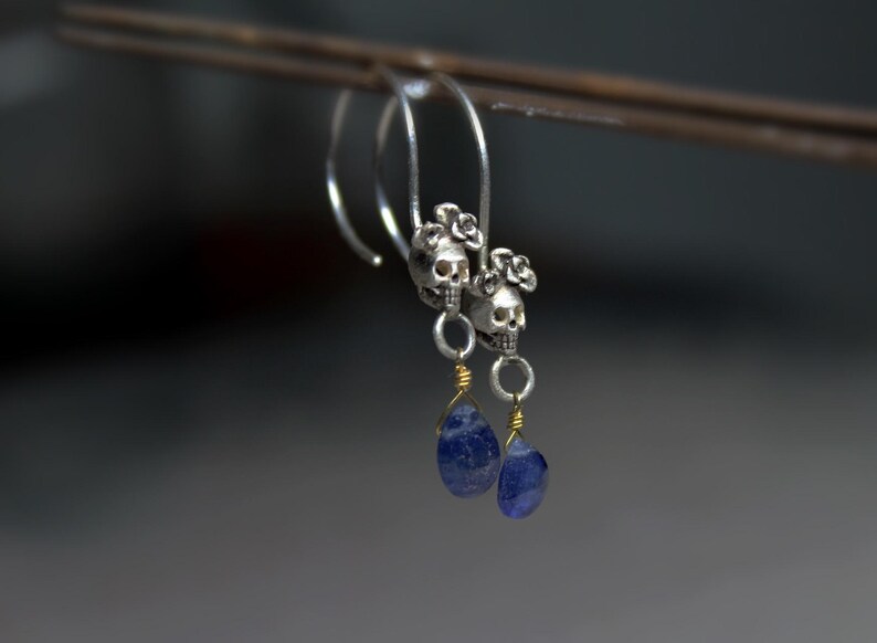 May include: A pair of silver skull earrings with blue gemstone drops. The skulls are adorned with small flower accents.