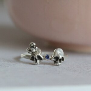 May include: A silver ring with two skull designs and a blue gemstone in the center.