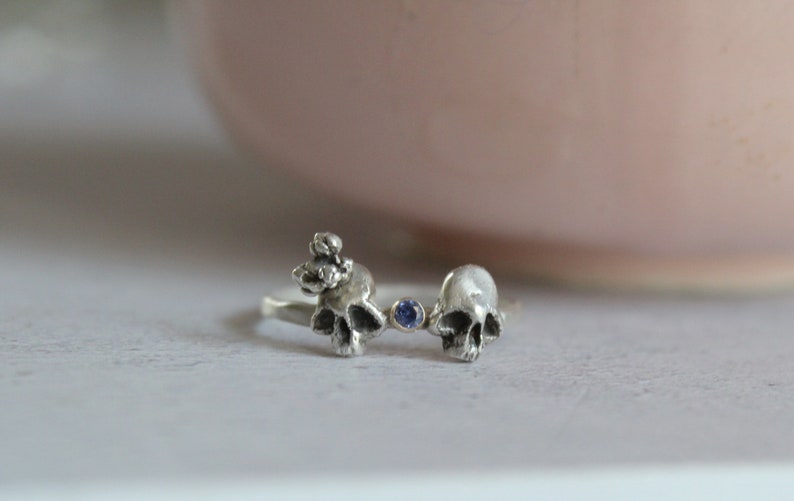 May include: A silver ring with two skull designs and a blue gemstone in the center.