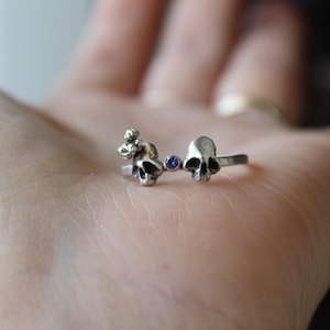 May include: A silver ring with two skull charms and a blue gemstone in between. The ring is on a person's hand.