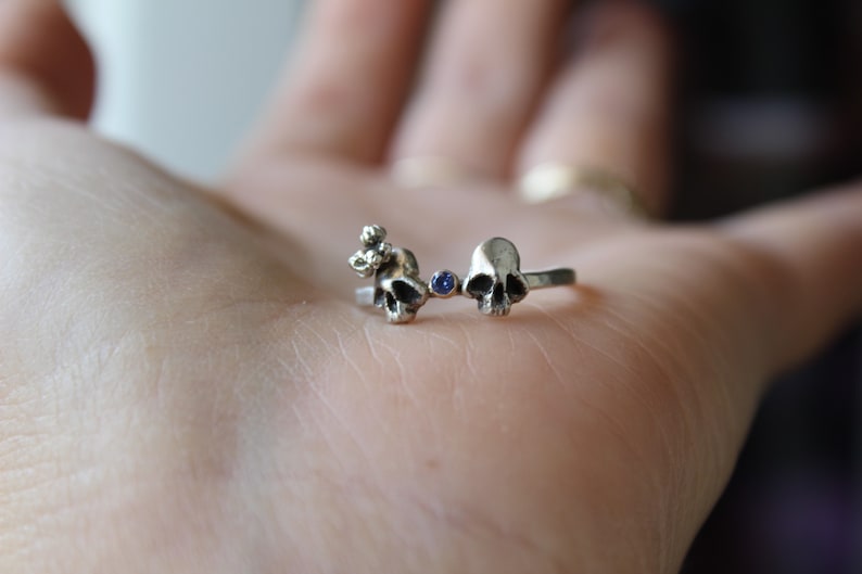 May include: A silver ring with two skull charms and a blue gemstone in between. The ring is on a person's hand.