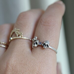May include: A silver ring with two small skull charms and a purple gemstone. The ring is on a person's finger.