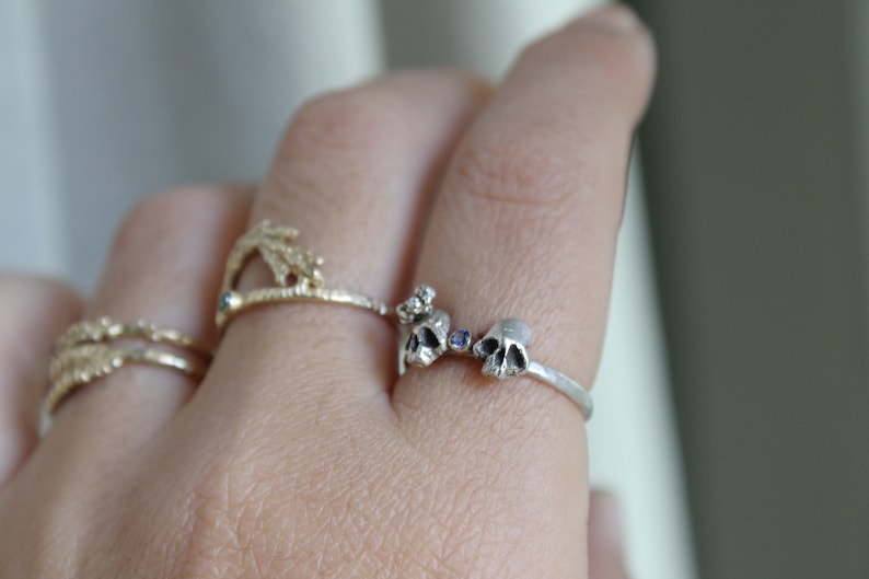 May include: A silver ring with two small skull charms and a purple gemstone. The ring is on a person's finger.