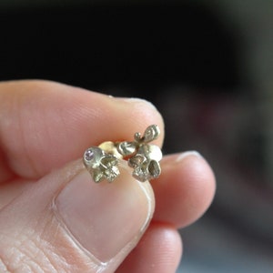 Peut inclure: Une paire de boucles d'oreilles en forme de cr&acirc;ne en or avec de petites pierres pr&eacute;cieuses roses. Les boucles d'oreilles sont tenues dans une main.