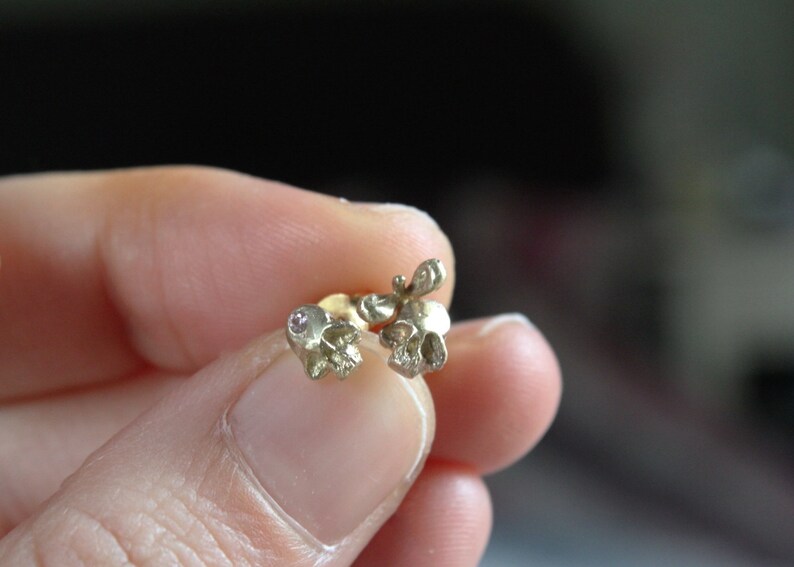 Peut inclure: Une paire de boucles d'oreilles en forme de cr&acirc;ne en or avec de petites pierres pr&eacute;cieuses roses. Les boucles d'oreilles sont tenues dans une main.