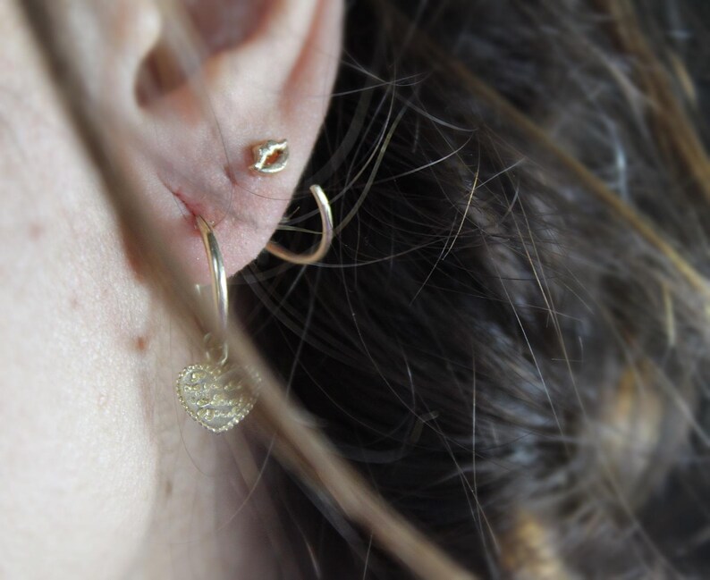 May include: Gold hoop earrings with a small heart charm dangling from one earring. The heart charm is covered in small, sparkling stones.