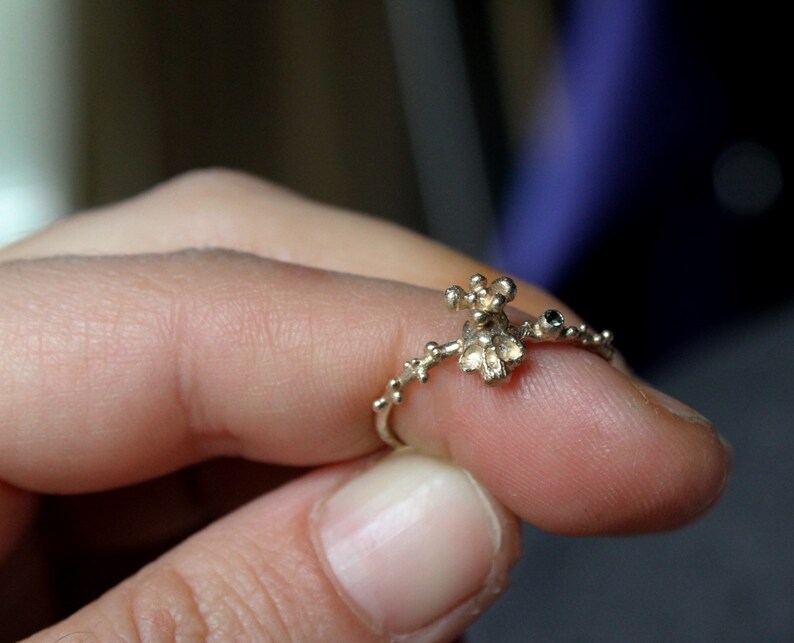 May include: A delicate silver ring with a small, intricate design featuring a cluster of tiny, rounded shapes. The ring is adorned with a single, dark blue gemstone.