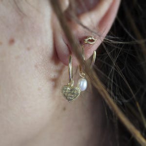 May include: A person wearing gold hoop earrings with a heart charm and a pearl dangle earring.