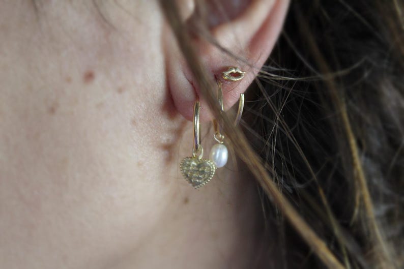 May include: A person wearing gold hoop earrings with a heart charm and a pearl dangle earring.