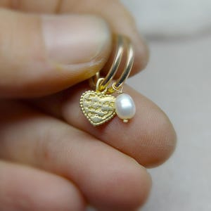 May include: Gold hoop earrings with a small heart charm and a pearl dangle. The heart charm has a textured surface and the pearl is white.