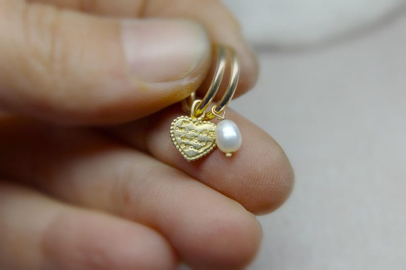 May include: Gold hoop earrings with a small heart charm and a pearl dangle. The heart charm has a textured surface and the pearl is white.