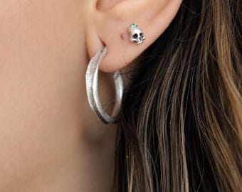 May include: A silver hoop earring with a textured surface and a small silver stud earring shaped like a skull with a tiny green gemstone set in the forehead. Both pieces are worn in the earlobe, showcasing a metallic finish and detailed design.