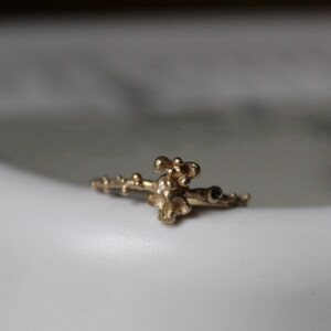 May include: A gold ring with a delicate, organic design featuring a cluster of small, rounded shapes. The ring has a textured surface and a small black stone.