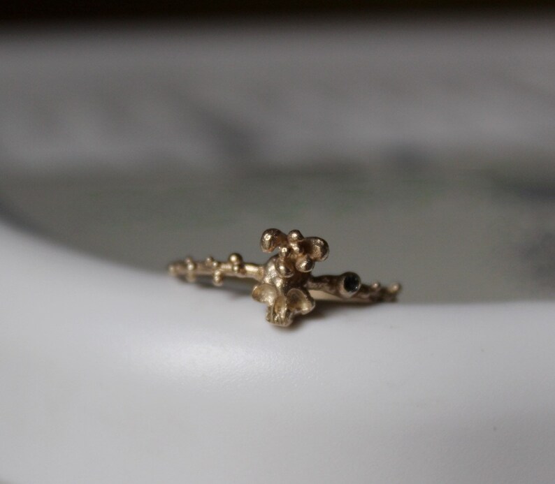 May include: A gold ring with a delicate, organic design featuring a cluster of small, rounded shapes. The ring has a textured surface and a small black stone.