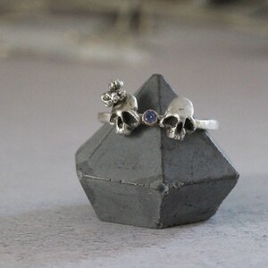 May include: A silver ring with three skull charms and a small blue gemstone. The ring is displayed on a gray concrete stand.