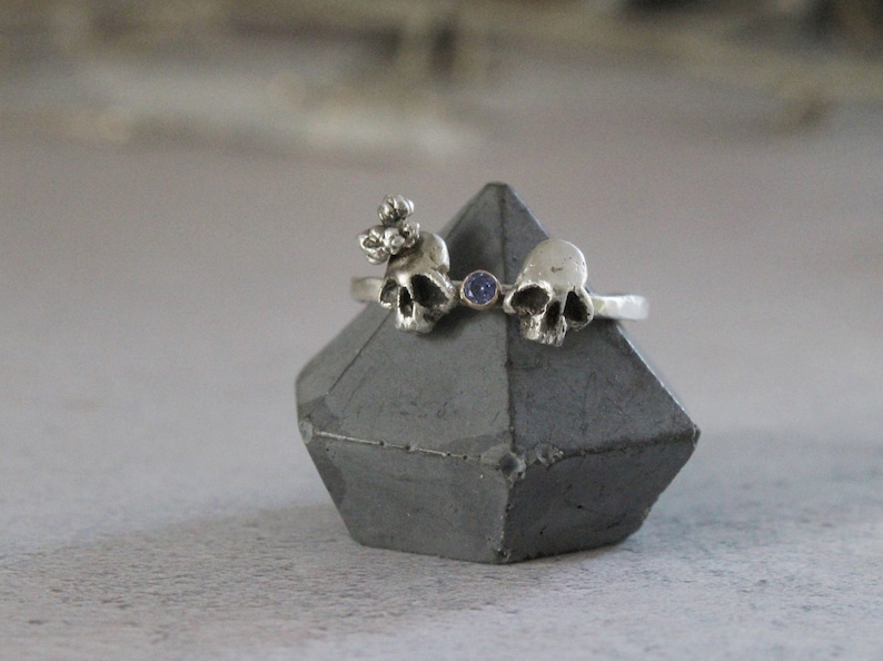 May include: A silver ring with three skull charms and a small blue gemstone. The ring is displayed on a gray concrete stand.