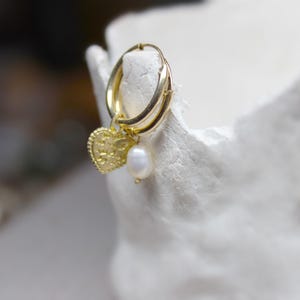 May include: Gold hoop earrings with a small heart charm and a pearl dangle. The earrings are on a white surface.
