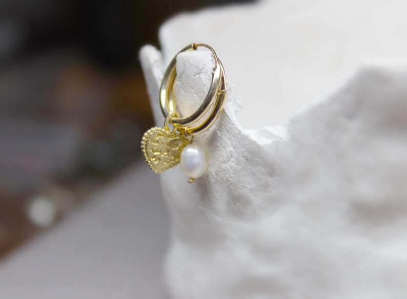 May include: Gold hoop earrings with a small heart charm and a pearl dangle. The earrings are on a white surface.