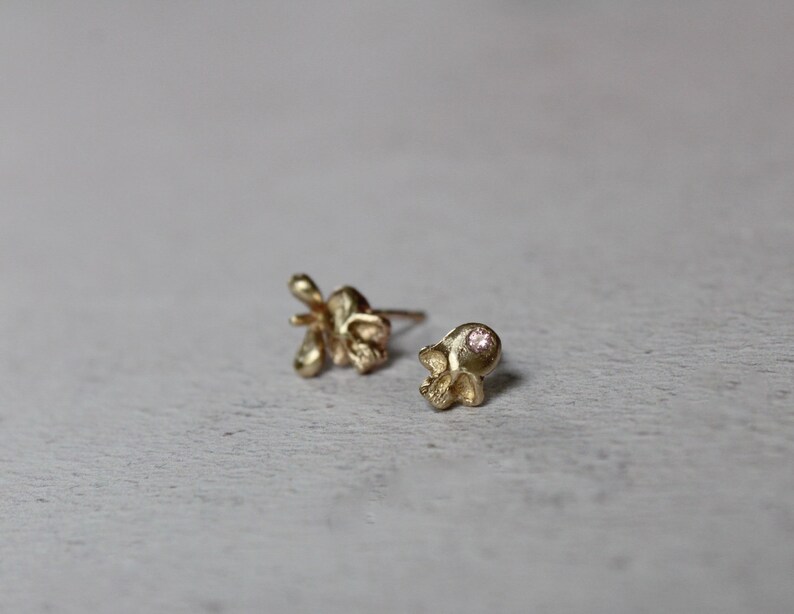 Peut inclure: Une paire de boucles d'oreilles en or, l'une en forme de fleur et l'autre en forme de cr&acirc;ne. La boucle d'oreille en forme de cr&acirc;ne a une petite pierre pr&eacute;cieuse rose dans l'orbite de l'&oelig;il.